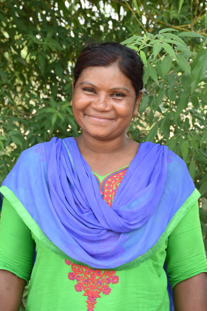 A smiling Munni Keshariyar, wearing a colourful dress, stands against a backdrop of leaves.
