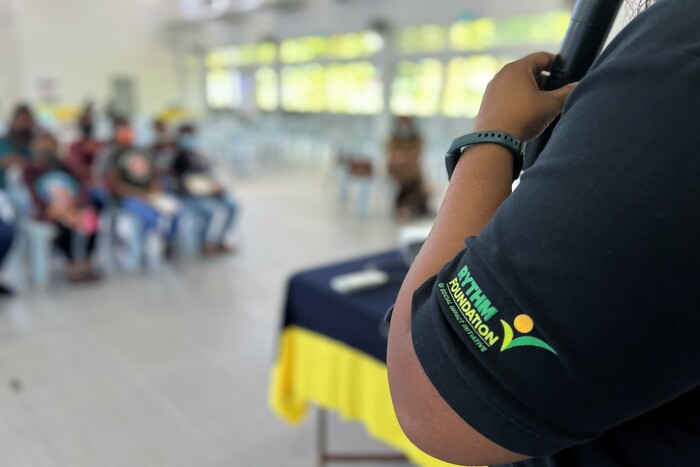 A close-up of a person's arm with 'RYTHM Foundation' written on the sleeve, while the background shows an out-of-focus group of people seated.