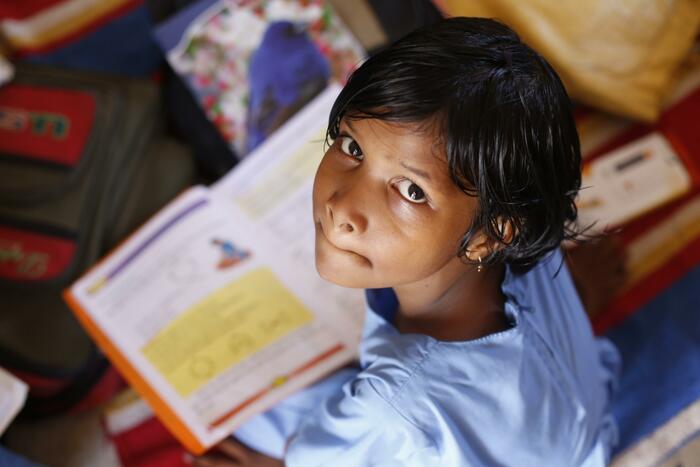 A young girl in a blue dress looking up from her textbook.