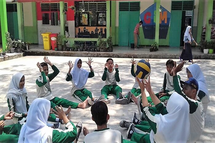 A group of students, wearing uniforms, sitting in a circle outside a colourful school building, playing volleyball.