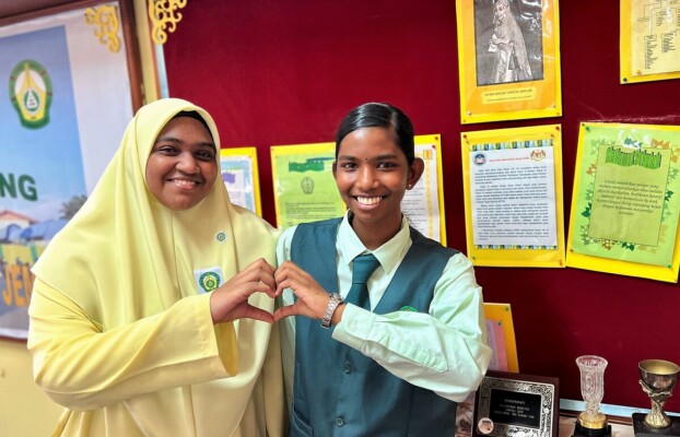 Two girls in school uniforms make a mini heart gesture with their fingers.