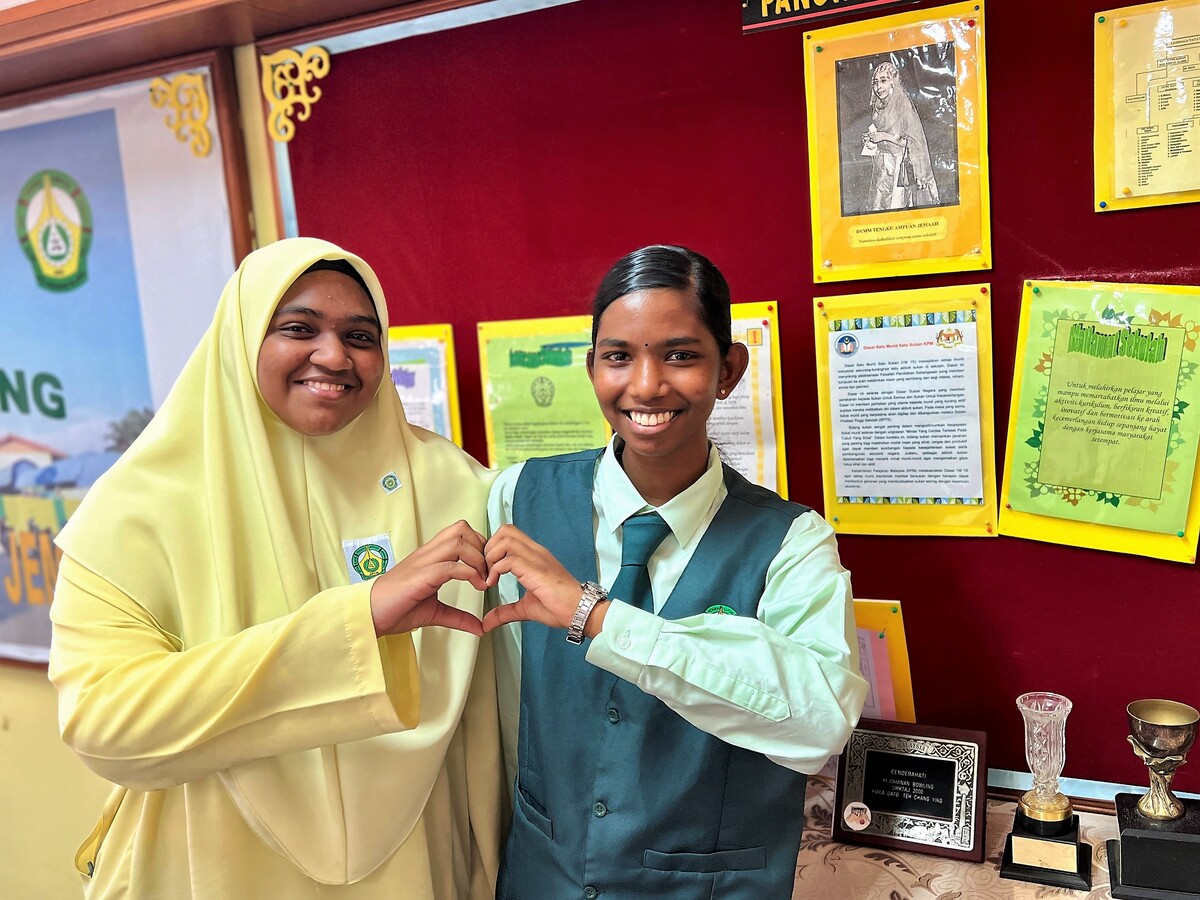 Two girls in school uniforms make a mini heart gesture with their fingers.