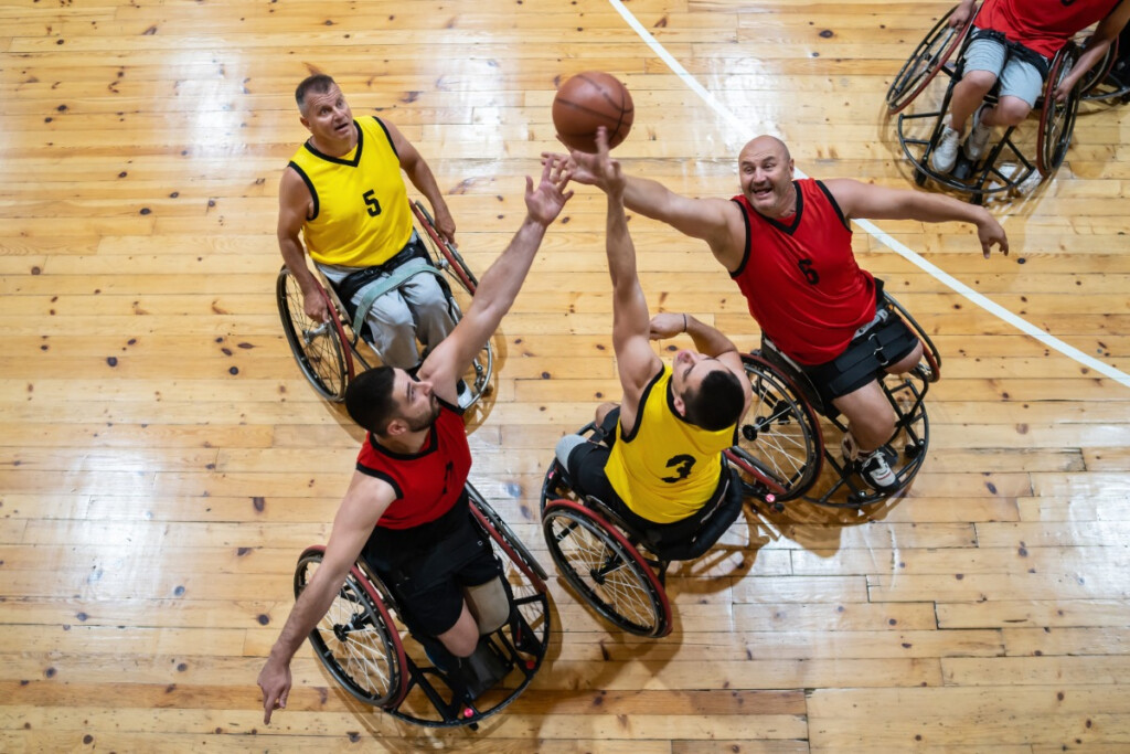 Two teams of athletes with disabilities in wheelchairs compete in a game of wheelchair basketball on a wooden court.