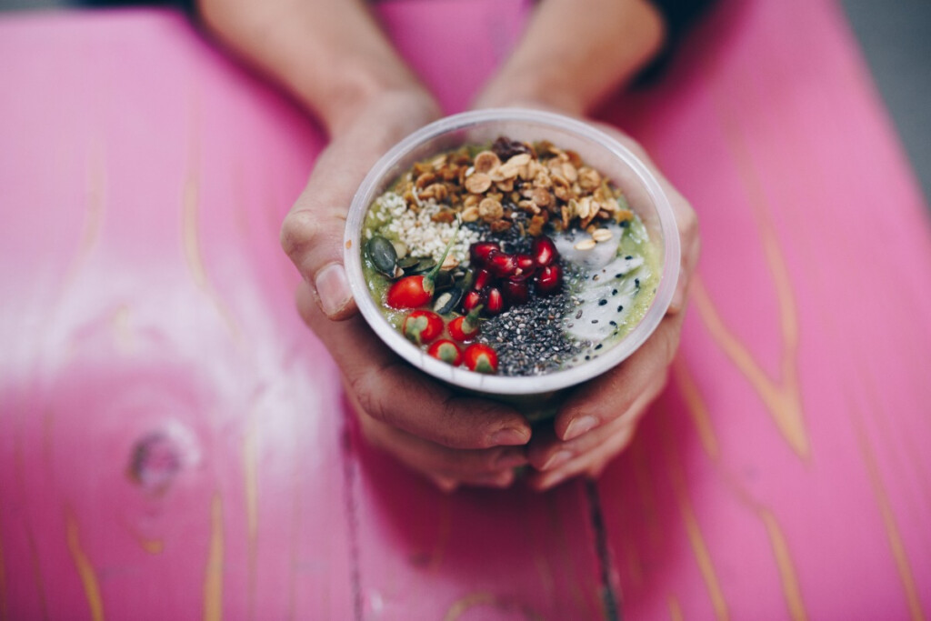 A person holding a bowl of healthy food with seeds, pomegranate, and granola on a pink table background.