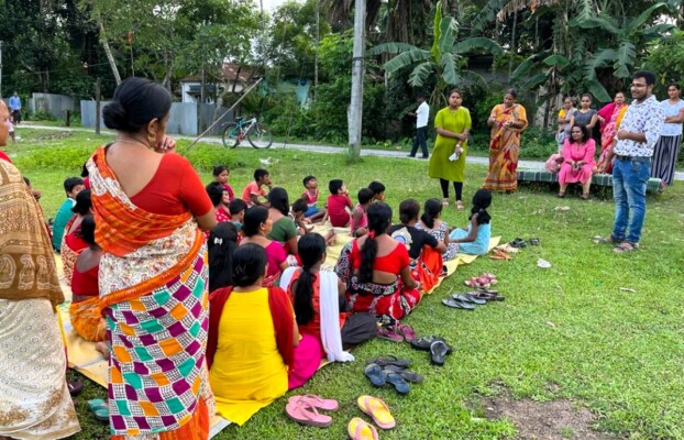A group of women and girls sitting on the ground outdoors in North Bengal, India, attentively listening to a literacy talk.
