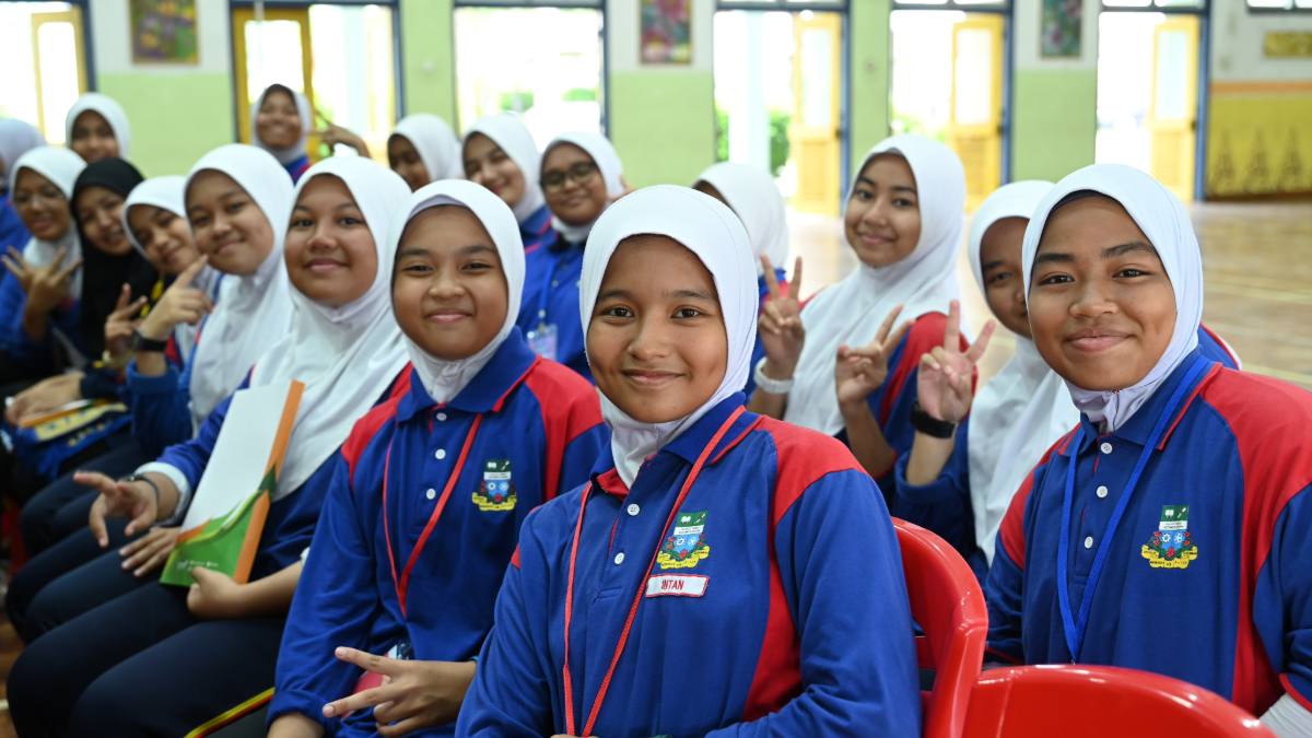 A group of cheerful young girls in school uniforms, smiling and making peace signs during an educational program in a school hall.