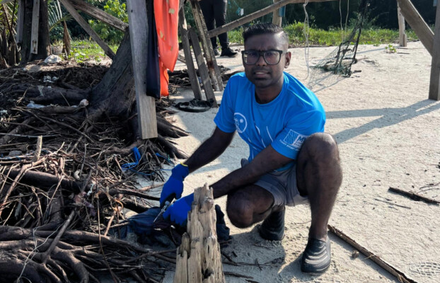 Nimal crouches on a sandy beach, removing debris near a makeshift shelter during a clean-up effort.
