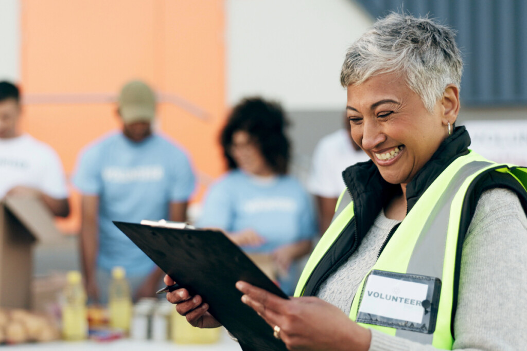 A smiling volunteer reviewing a clipboard at a community event, with other volunteers working in the background.
